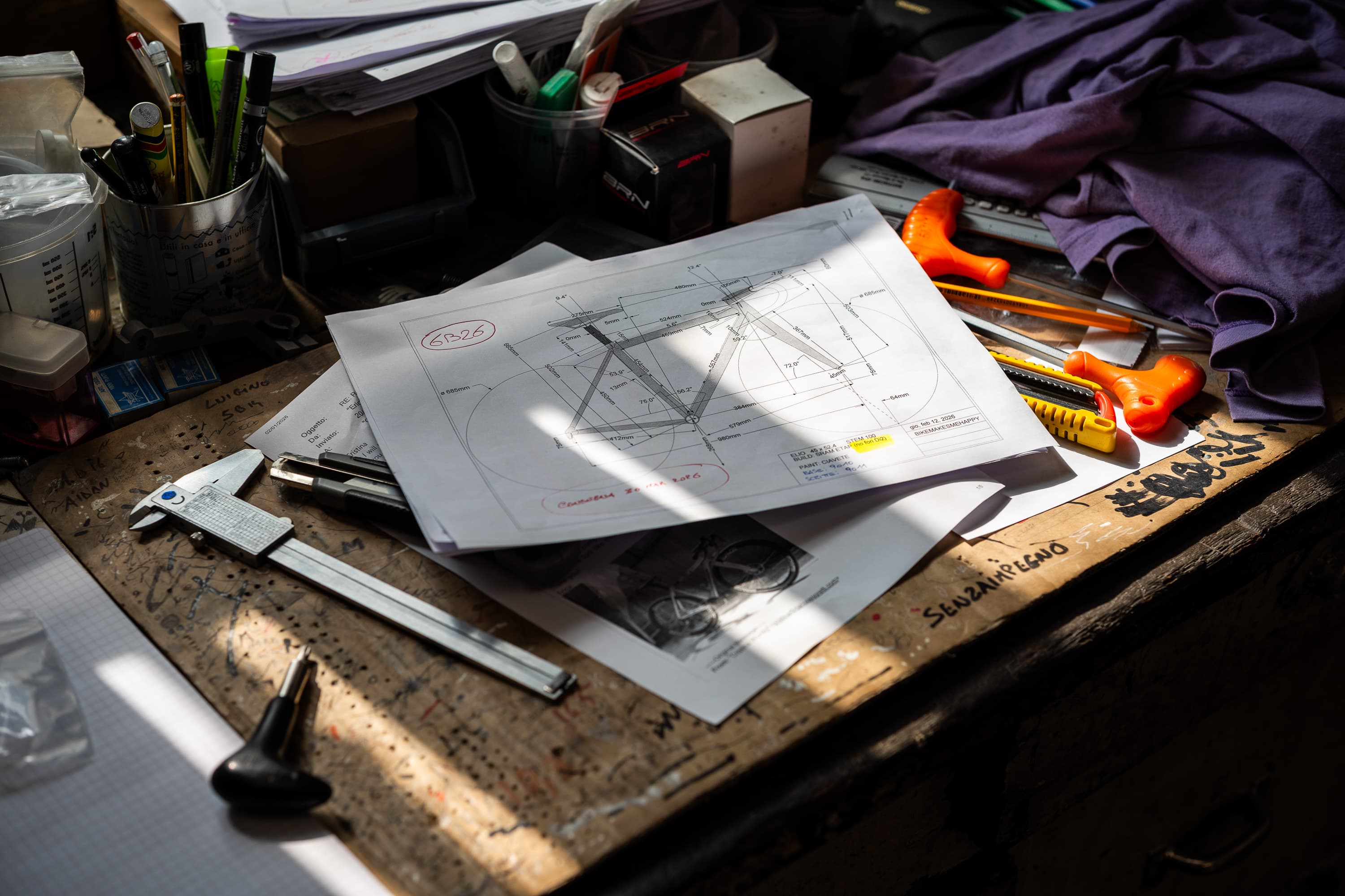 A bicycle frame's technical geometry drawing resting on a graffiti-covered workshop bench, surrounded by calipers, craft knives, pens and a steel rule