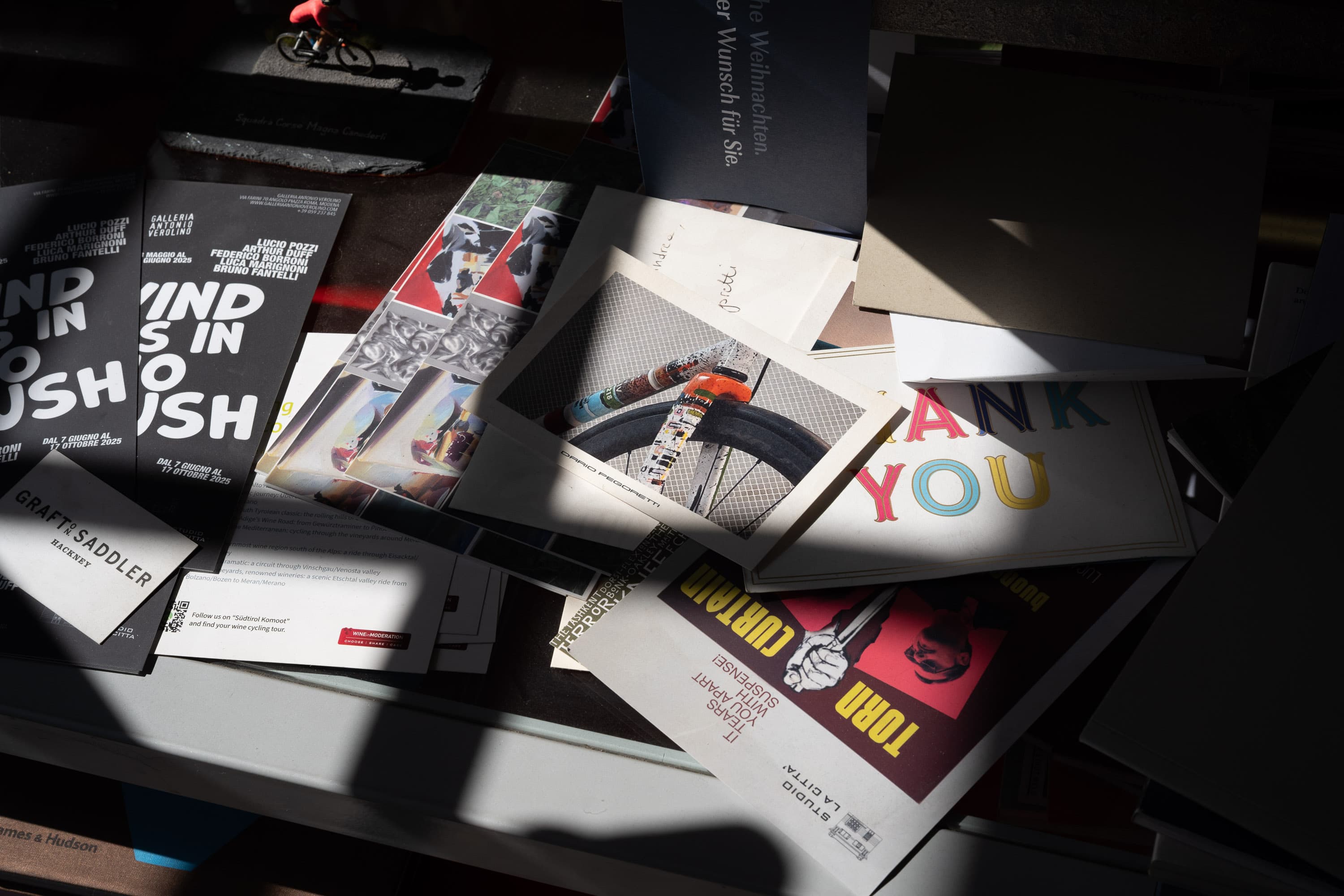 A studio desk in hard sunlight, layered with printed matter — gallery flyers, a thank-you card, and a Dario Pegoretti postcard of a painted fork crown