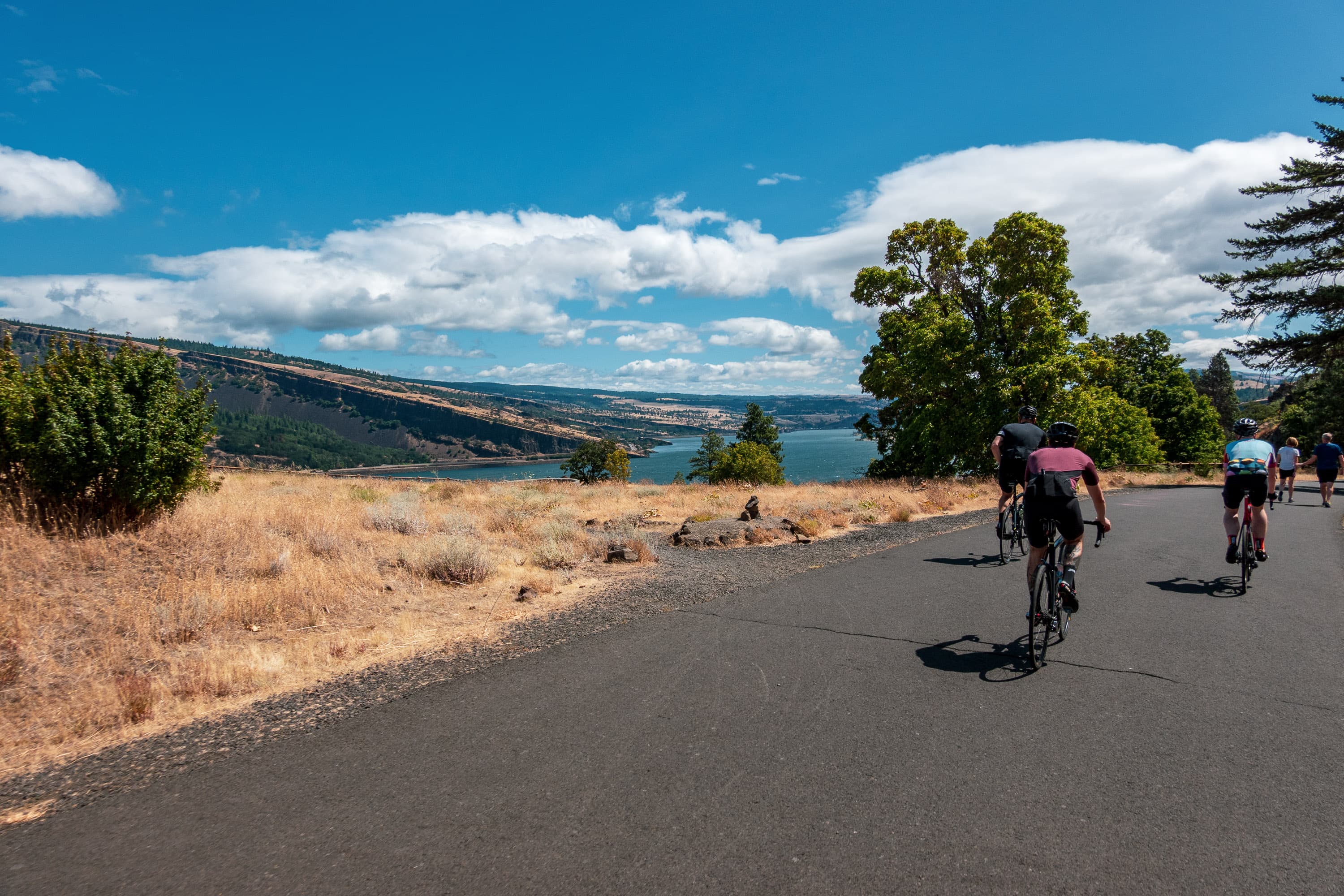 Cyclists riding a smooth path above a wide blue river gorge under summer clouds, walkers a little further on